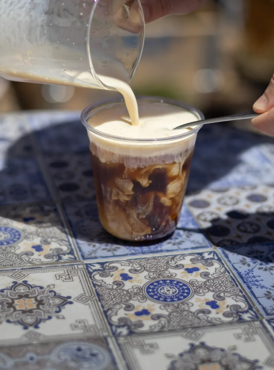 Iced coffee being topped with cold cream, on a blue and white tiled table in the sun